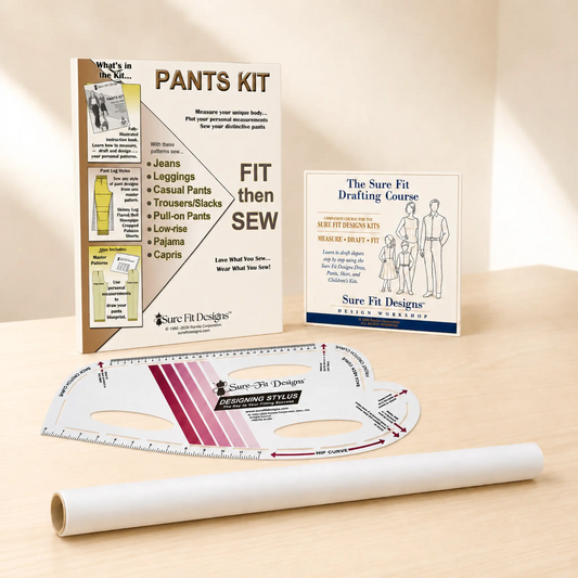 A sewing pattern kit set on a light wood surface with a warm beige background. The set includes a white pattern transfer sheet, a white roll of transfer paper, a metal ruler, and two printed instruction/booklet cards: one titled 'PANTS KIT' and the other 'The Sure Fit Drafting Course'.