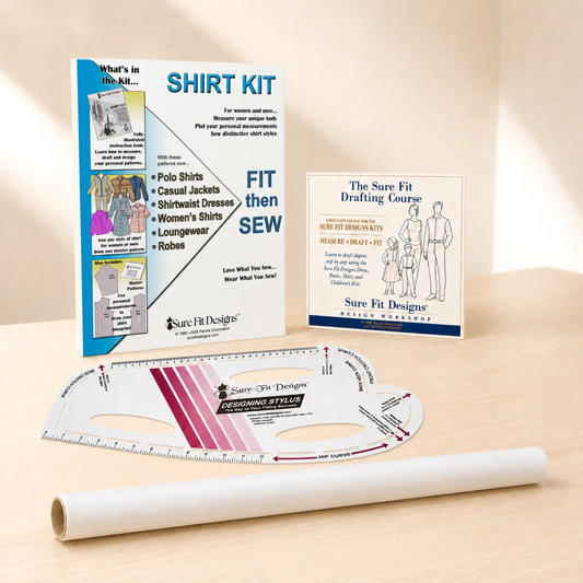 A sewing and garment-making kit displayed on a light wood surface, including a large instruction booklet, a drafting template ruler, a roll of white fabric, and a smaller course booklet. Visible branding includes Sure Fit Designs.
