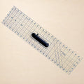 A clear acrylic rotary cutter with blue grid lines and a black handle, shown diagonally on a light beige background. The brand 'Made In Design' is printed on the handle.